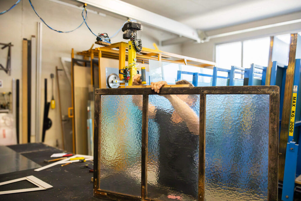 Handwerker in einer Glaserei hebt ein Fenster mit strukturiertem Ornamentglas und Holzrahmen mithilfe eines Vakuumhebers an. Im Hintergrund sind moderne Glasscheiben und Werkstatteinrichtung sichtbar.